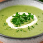 Eine Schale cremige grüne Kerbelsuppe, garniert mit einem Sahnekringel und frischem Kerbel, serviert in einer rustikalen Keramikschale auf einem Holztisch.